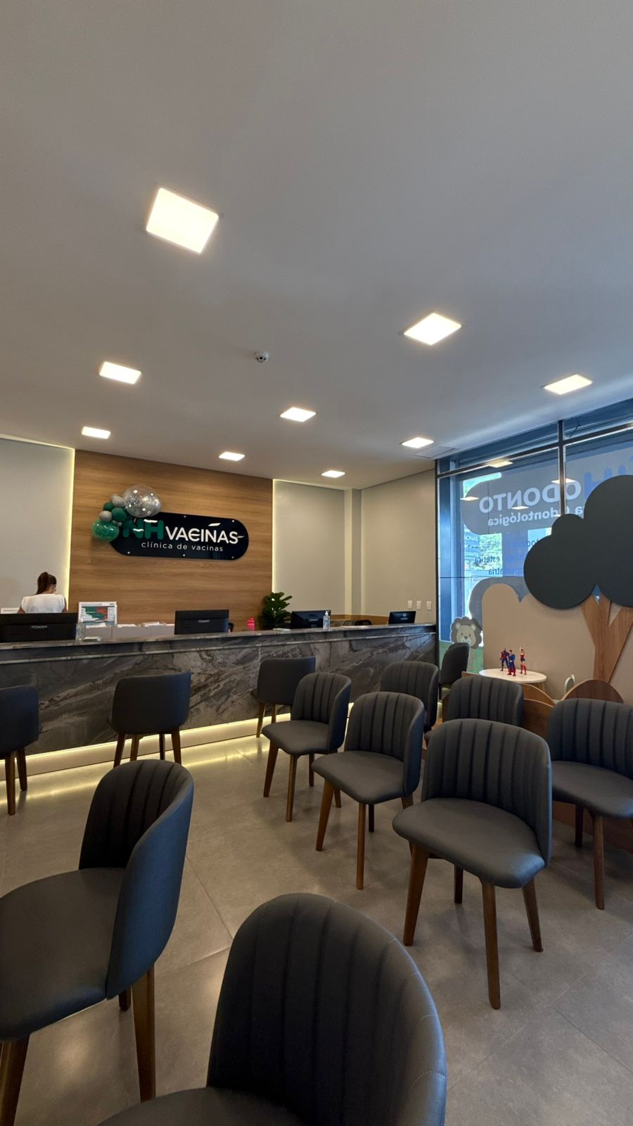 Recepção da NH Vacinas com logo, cadeiras cinza e brinquedoteca ao fundo