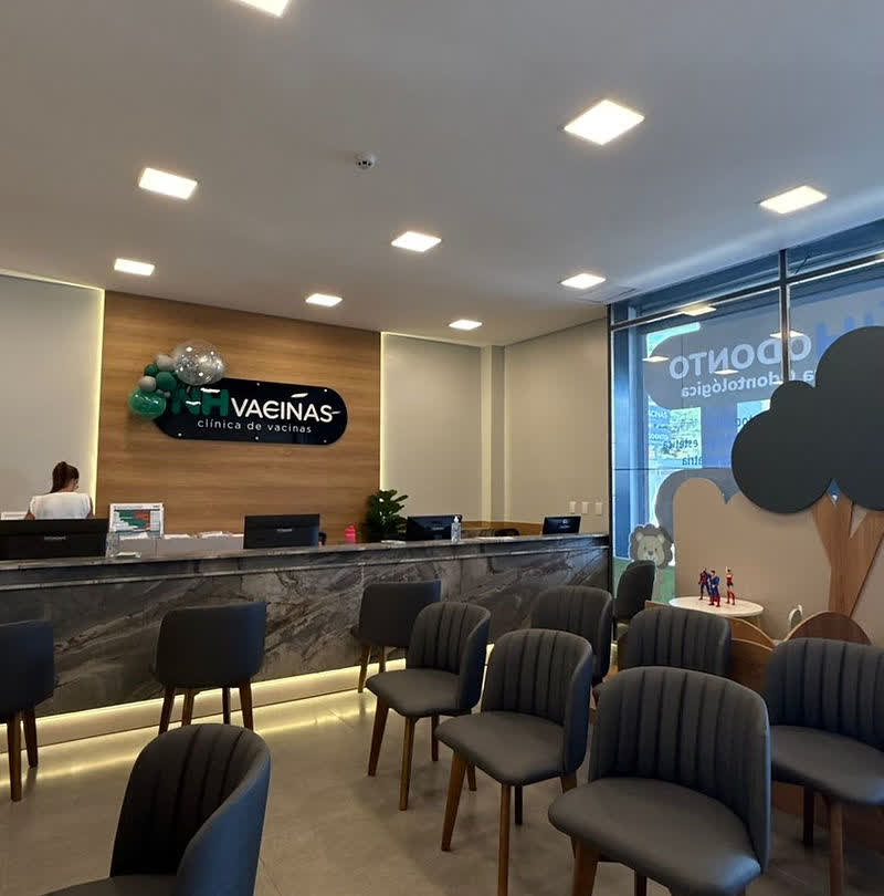 Recepção da NH Vacinas com balcão de madeira, logo NH VACINAS, cadeiras cinza e brinquedoteca